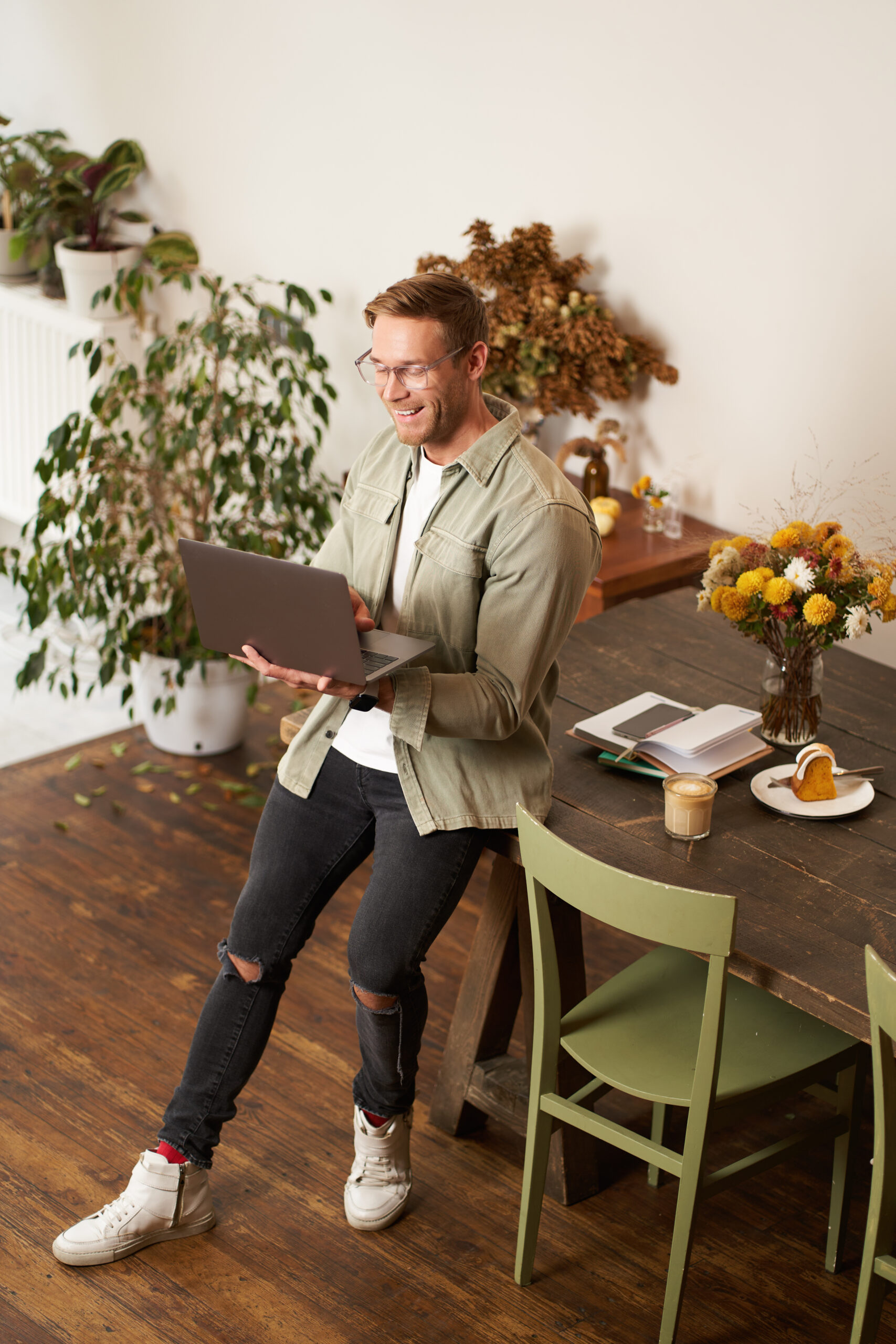 Vertical portrait of handsome happy man, manager or CEO of company, sitting on table and holding laptop, laughing, video chatting, has an online meeting with a team, standing in an office